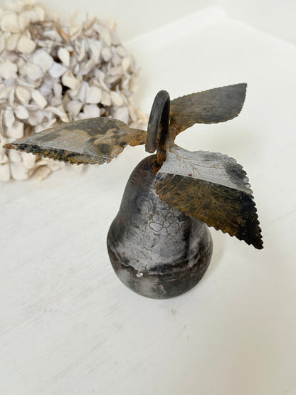 A rustic metal bird bell decoration with a distressed dark gray and brown patina, displayed on a white surface next to dried flowers.