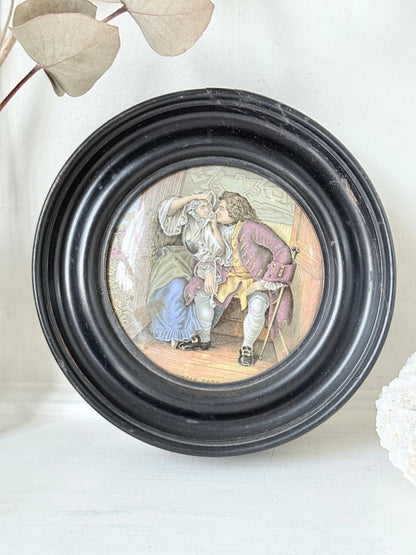 Antique Prattware pot lid in a black round frame with a detailed Victorian scene on a white surface.