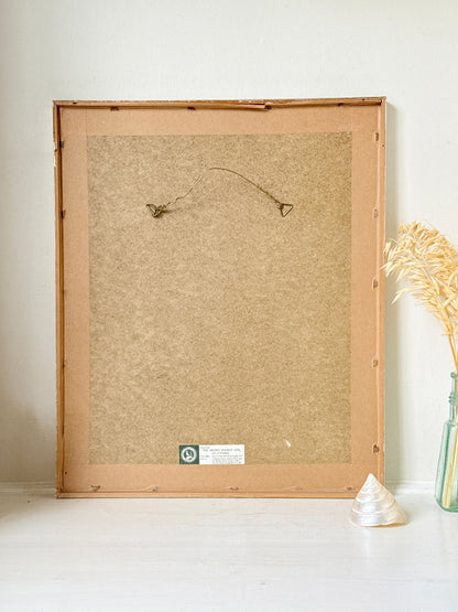 The back of a large square picture frame made of natural wood and cork, leaning against a white wall on a white floor, with a small decorative vase of dried grass and a seashell nearby.