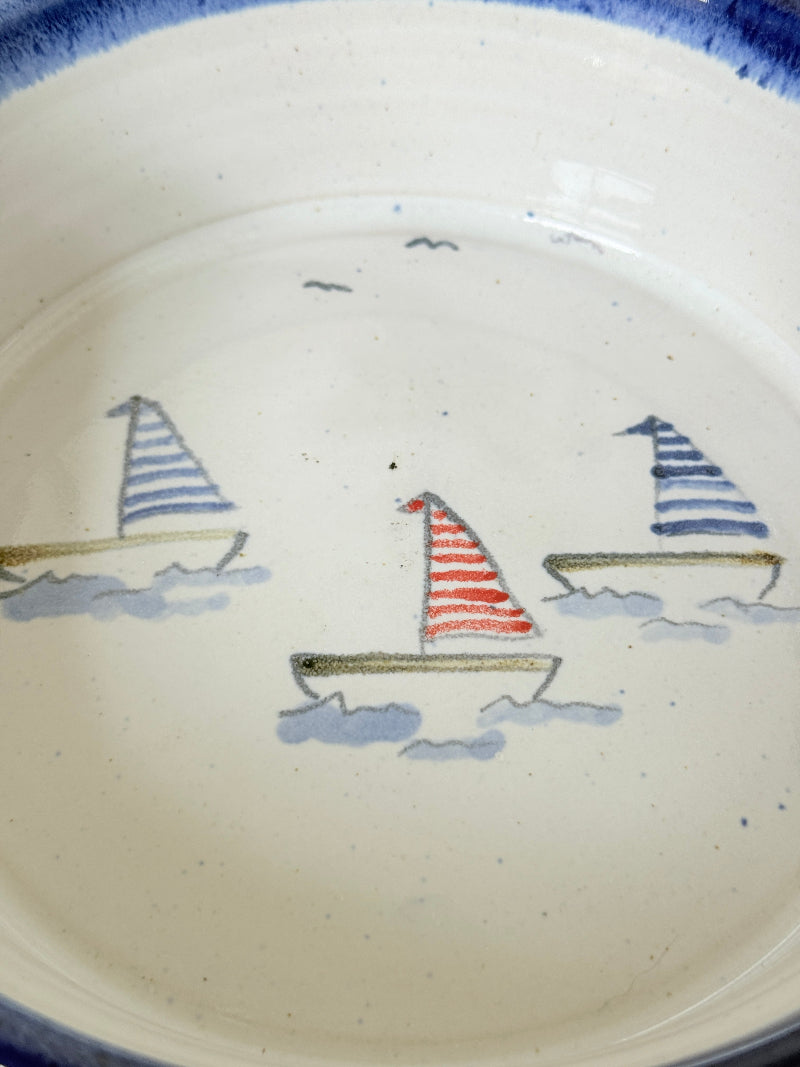 Close-up of a ceramic plate or bowl interior with a cream-colored glaze, decorated with three hand-painted sailboats in blue, red, and navy tones.