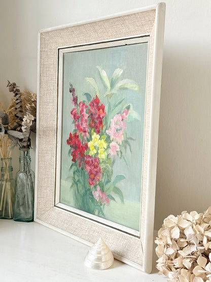 A framed floral painting with a textured cream linen-style frame, leaning against a wall on a tabletop with decorative glass bottles and dried flowers nearby.