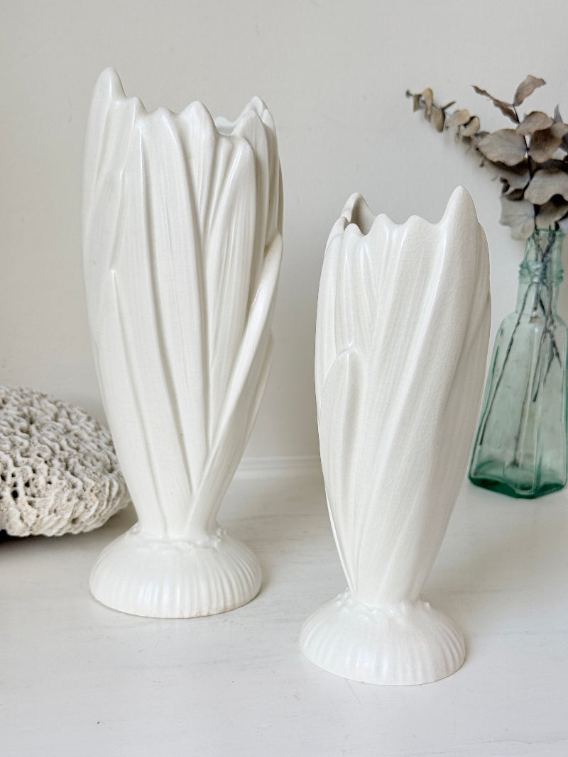 Two white ceramic tulip-shaped vases with fluted bases, displayed on a white surface against a light neutral background, with decorative props including a crocheted textile and a green glass vase with dried flowers.