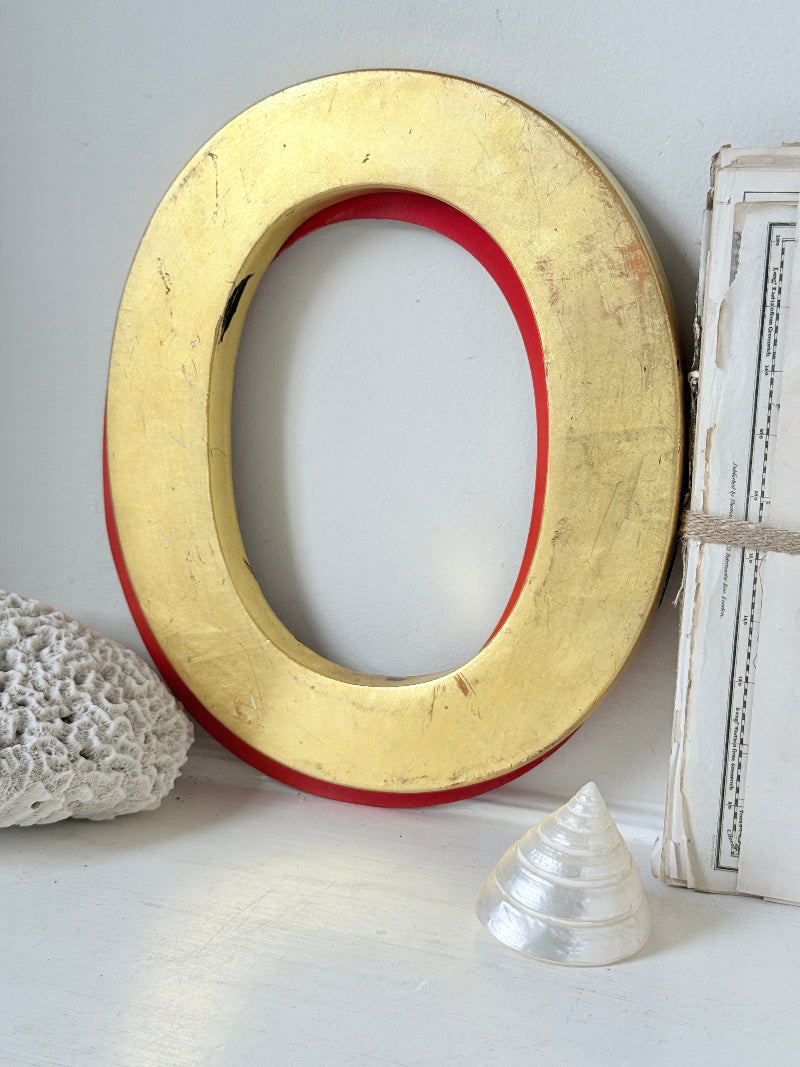 A large decorative letter O wall sign with a distressed gold finish and a red inner border, styled on a white surface with a white seashell, coral, and a vintage book.