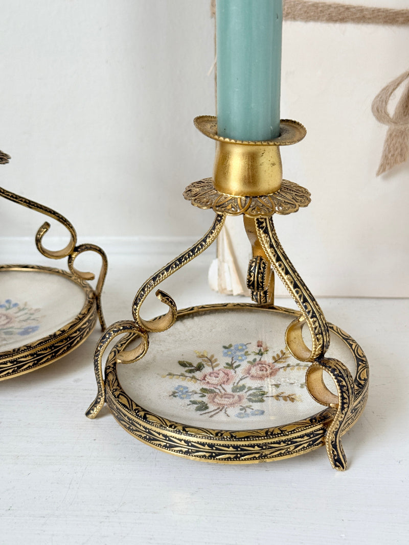 An ornate vintage-style gold metal candlestick holder with a floral porcelain base, holding a light blue candle, with another matching holder partially visible on the left.