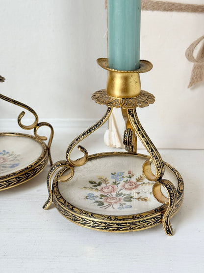 An ornate vintage-style gold metal candlestick holder with a floral porcelain base, holding a light blue candle, with another matching holder partially visible on the left.