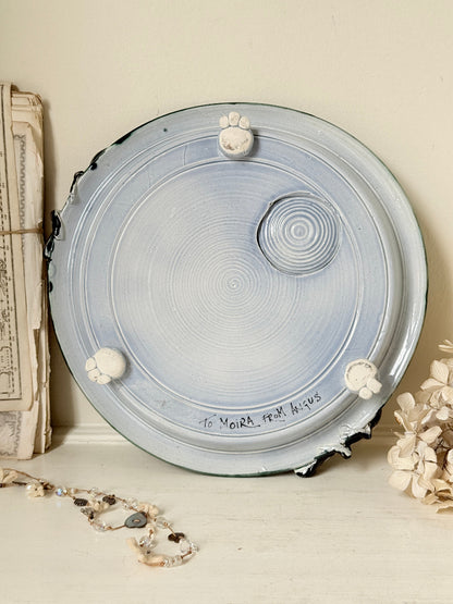 A handmade ceramic plate with a light blue and white spiral pattern, featuring three small white flower details and the text &