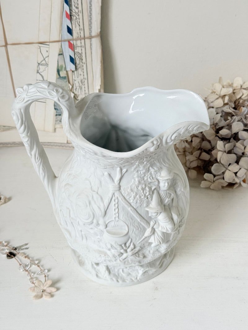 A white ceramic pitcher with intricate embossed floral and decorative patterns, set on a white surface with dried flowers and a vintage-style postcard in the background.