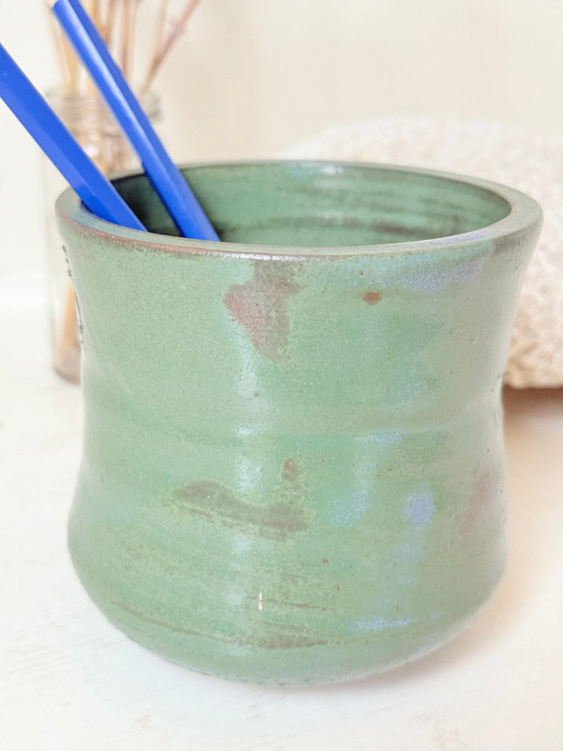Studio pottery pencil pot in green glaze, handmade on the wheel, photographed on a white surface with pencils inside.