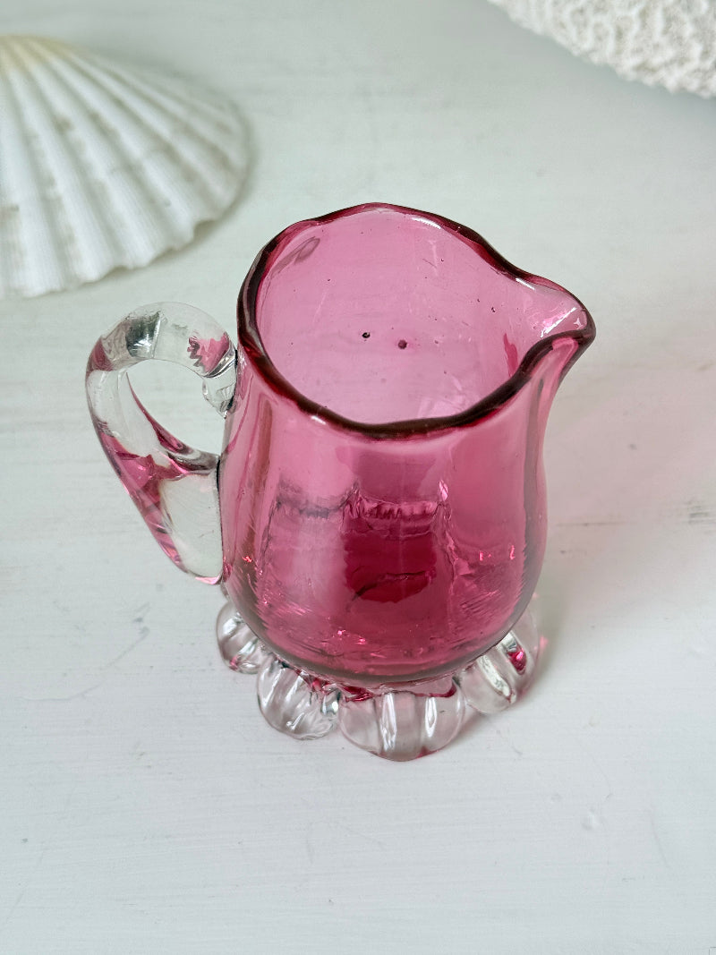 Antique cranberry glass cream jug with clear applied handle, crimped pedestal foot and deep pink hand blown glass body