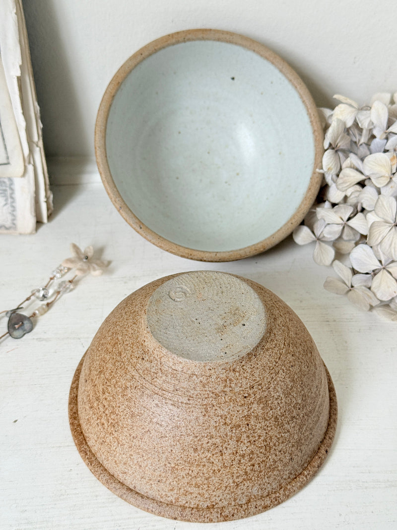 Two handmade ceramic bowls with speckled brown and beige glazes, one shown upside down and one standing upright, styled on a white wooden surface with dried flowers and vintage paper props.