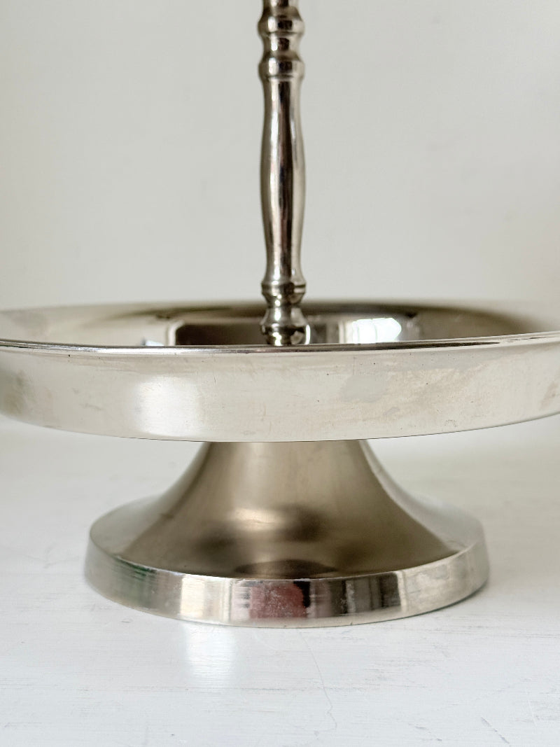 Two tier silver tone cake stand with polished reflective finish styled with green wrapped sweets on a white surface