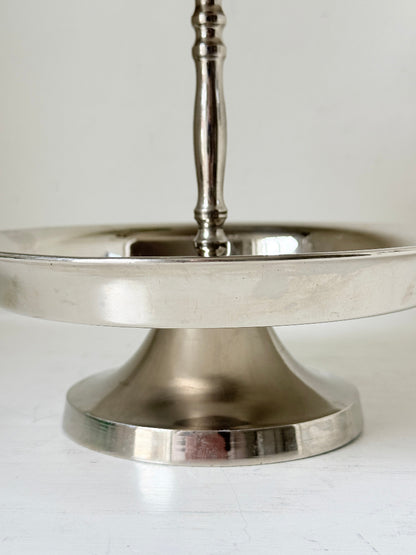 Two tier silver tone cake stand with polished reflective finish styled with green wrapped sweets on a white surface