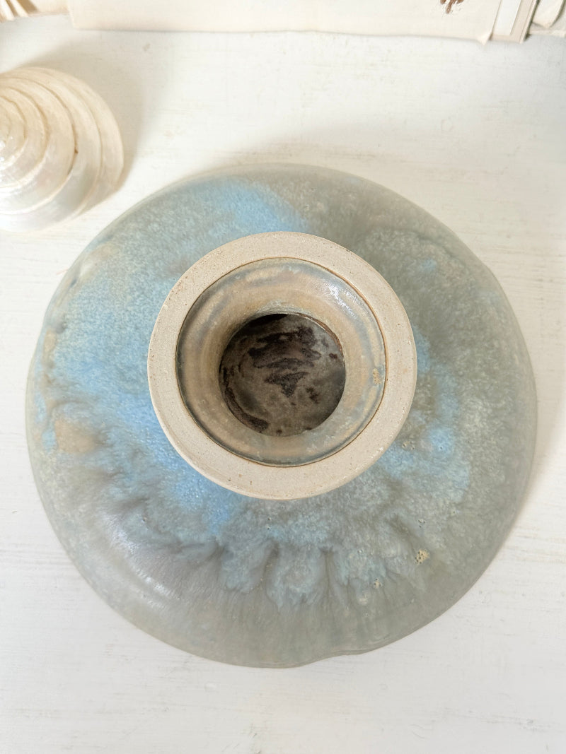 A round ceramic vase with a wide opening, featuring a light blue and gray glaze with a textured, marbled pattern, photographed from directly above on a white wooden surface.