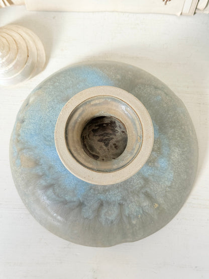 A round ceramic vase with a wide opening, featuring a light blue and gray glaze with a textured, marbled pattern, photographed from directly above on a white wooden surface.
