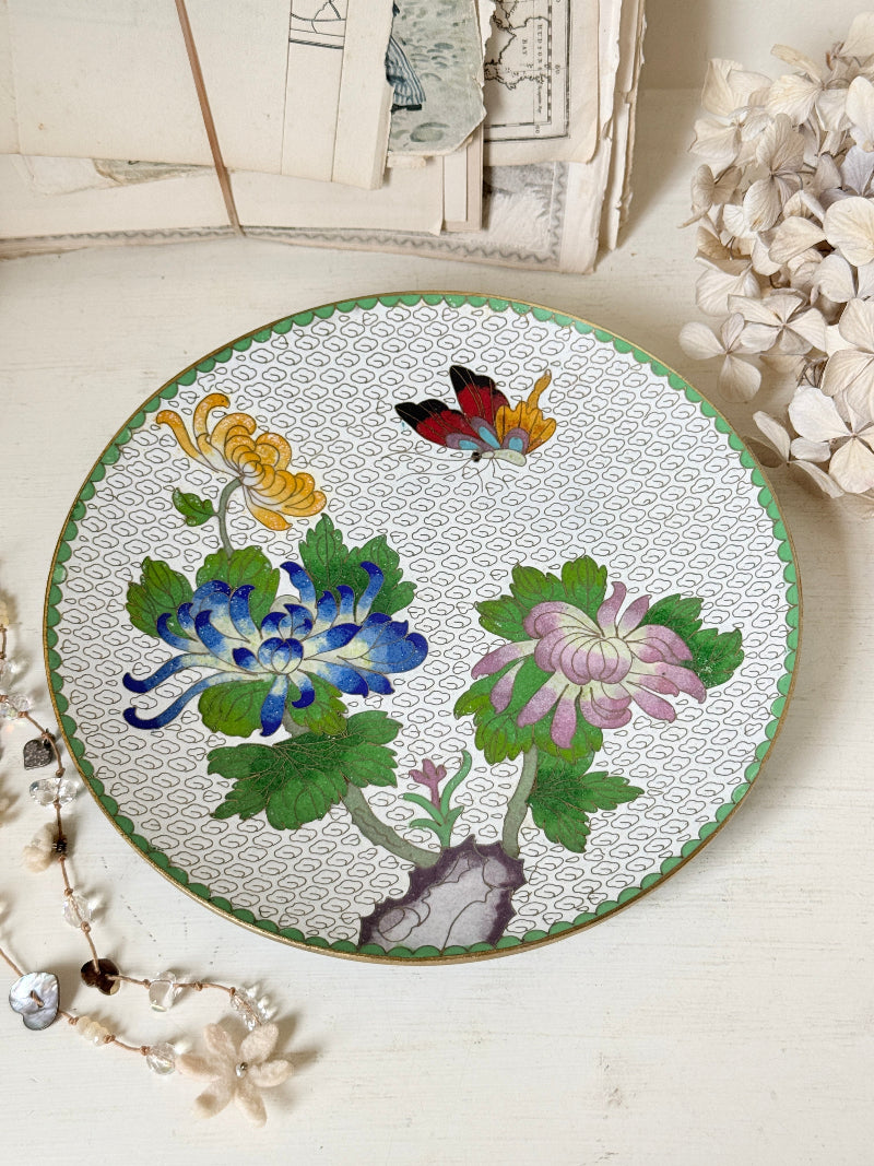 A round ceramic plate with a hand-painted floral design featuring blue, pink, and yellow chrysanthemums, green leaves, and a butterfly on a white patterned background, styled on a light wooden surface with dried flowers and vintage papers.