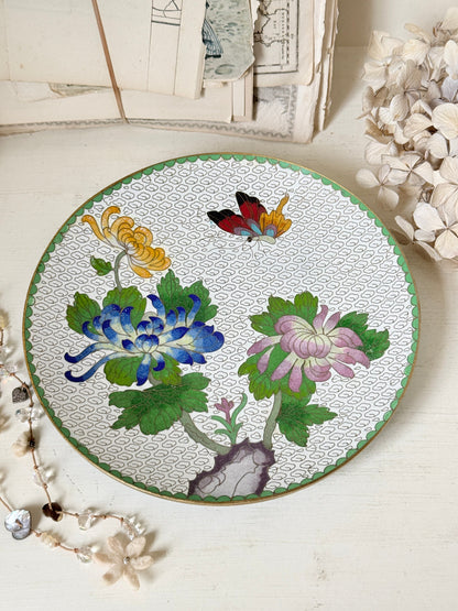 A round ceramic plate with a hand-painted floral design featuring blue, pink, and yellow chrysanthemums, green leaves, and a butterfly on a white patterned background, styled on a light wooden surface with dried flowers and vintage papers.