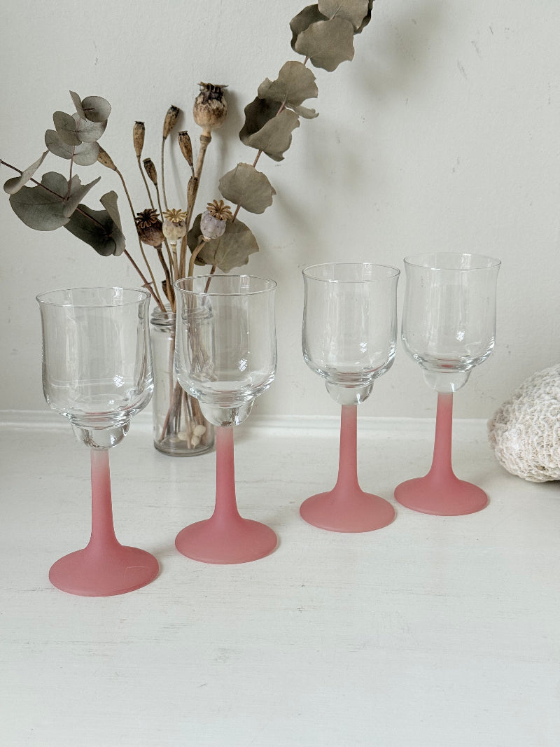 Set of four small vintage wine glasses with clear bowls and pink stems photographed on a white surface.
