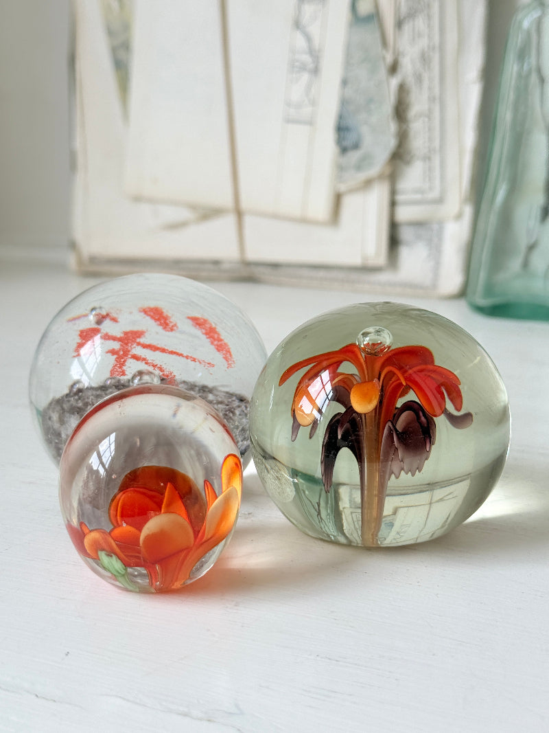 Trio of orange flower glass paperweights with green stems and controlled bubbles, photographed on a white surface