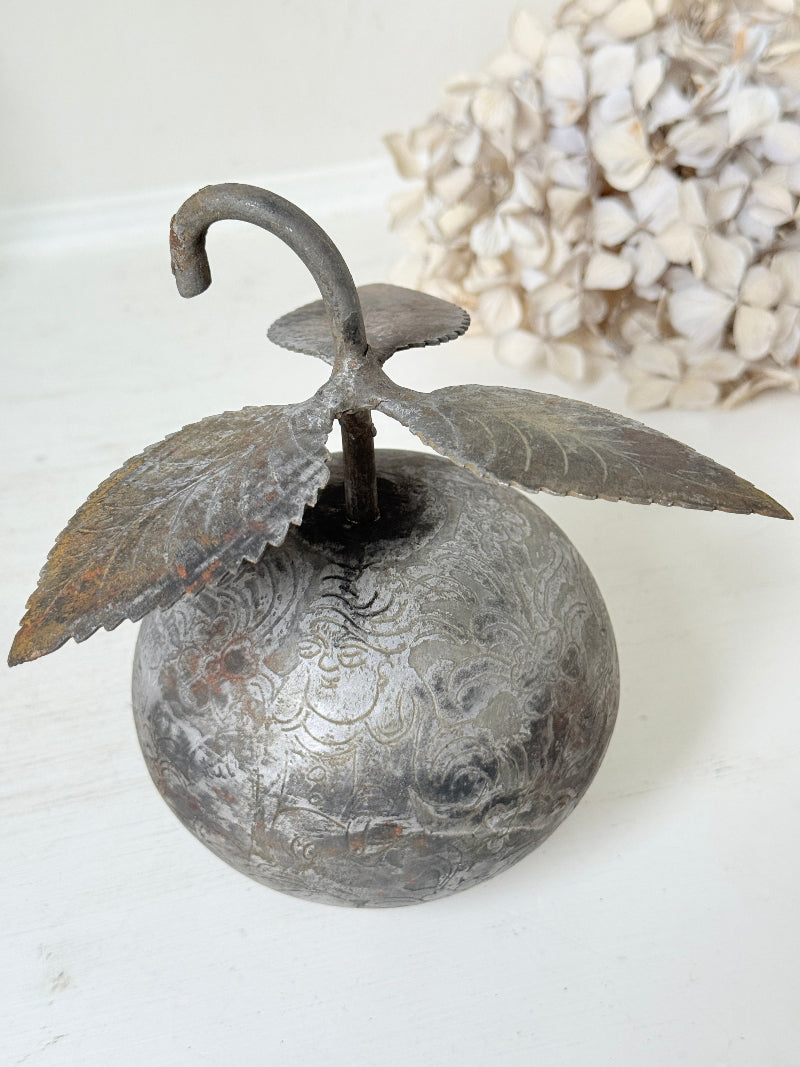 A decorative metal fruit-shaped object with a curved hook handle and leaf-like wings, shown on a white surface with a blurred cluster of dried flowers in the background.