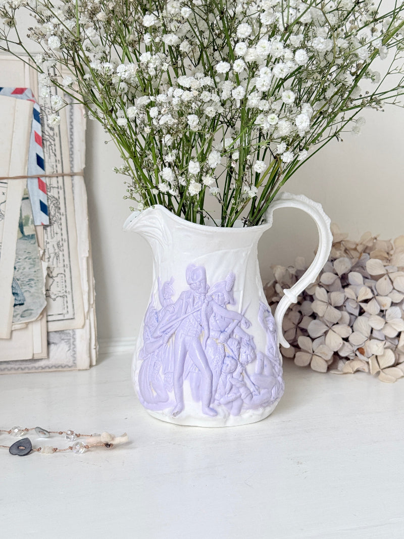 A white ceramic pitcher vase with embossed figures in pale lavender, filled with white baby&