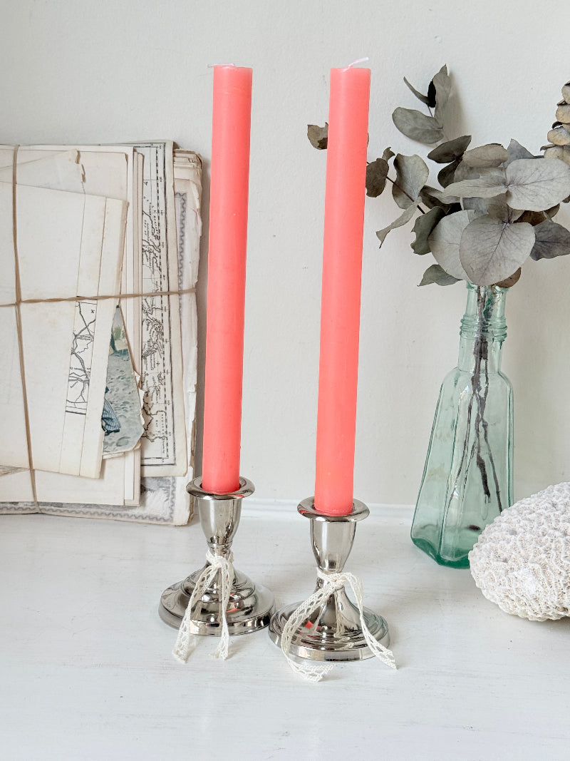 Pair of silver tone candlesticks with urn sconces and round stepped bases holding coral candles, photographed on a white surface