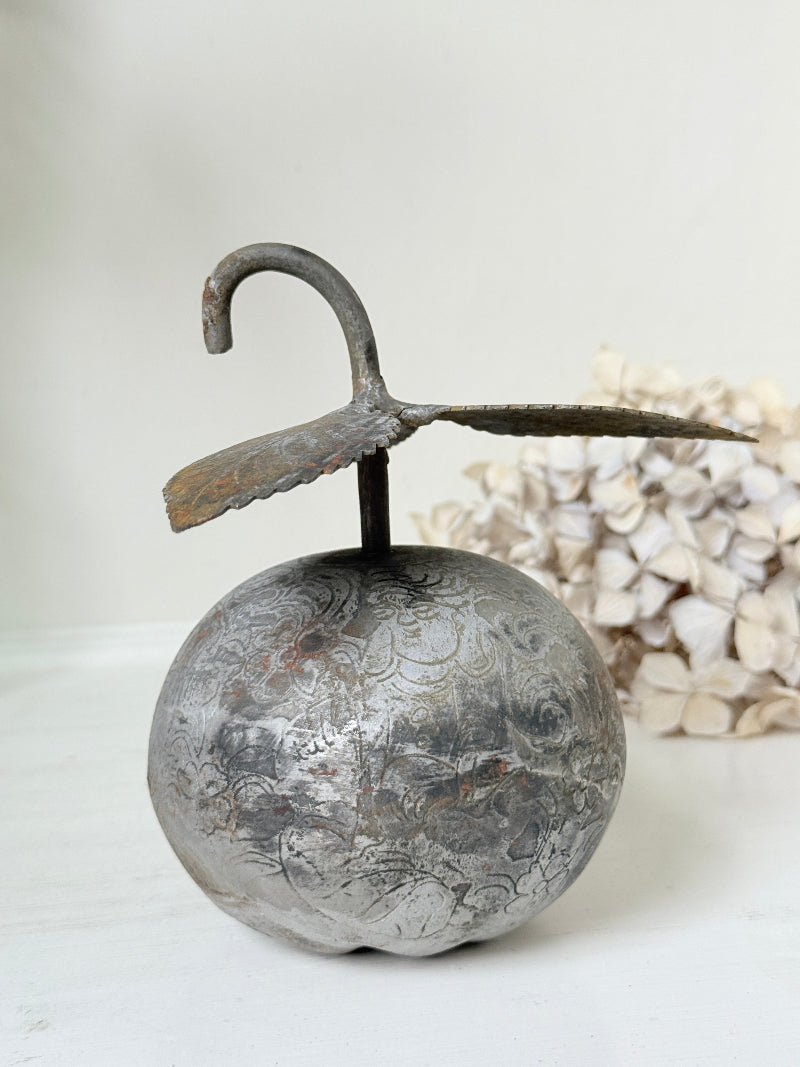 A rustic metal decorative object shaped like a round silver-toned fruit or orb with a curved hook-like stem, displayed on a light surface with a soft neutral background and dried flowers behind it.