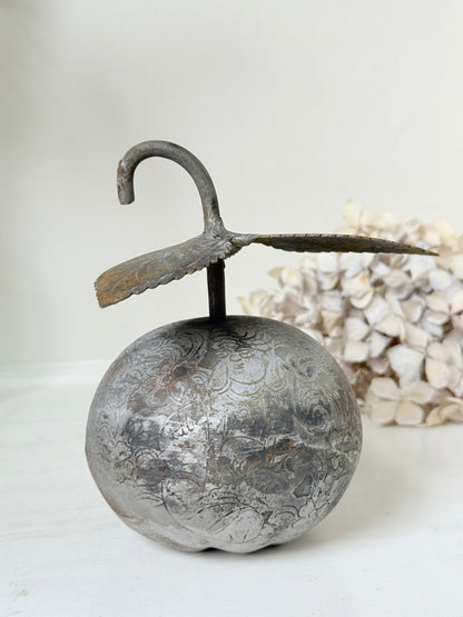 A rustic metal decorative object shaped like a round silver-toned fruit or orb with a curved hook-like stem, displayed on a light surface with a soft neutral background and dried flowers behind it.