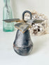 A vintage-style metal bird-shaped hanging ornament or decorative sculpture with a dark patinated surface, rusted brown highlights, and a curved top loop, photographed on a white surface with a blurred green glass bottle and dried flowers in the background.