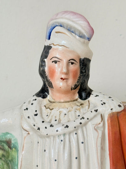 A close-up of a vintage ceramic figurine depicting a man in historical dress, featuring a speckled cream dress, black hair, and a pink and blue hat against a plain light background.