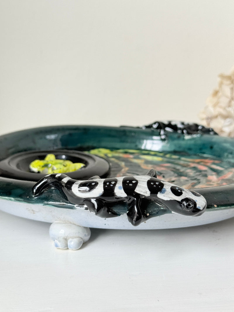 A handmade ceramic table decor piece featuring a black and white spotted lizard figure perched on a shallow oval dish with a dark green glazed interior and small white feet.