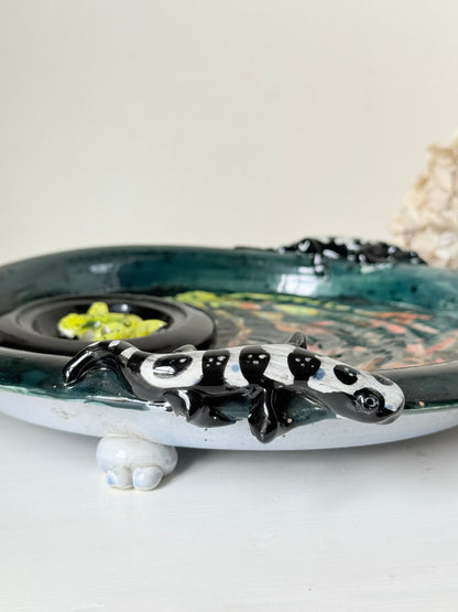 A handmade ceramic table decor piece featuring a black and white spotted lizard figure perched on a shallow oval dish with a dark green glazed interior and small white feet.