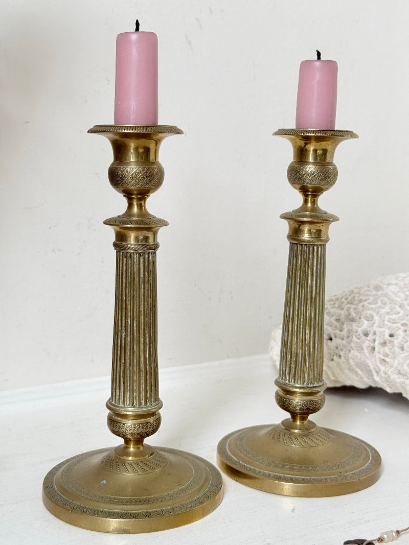 Pair of antique brass Empire style candlesticks photographed on a white surface with fluted columns and circular bases holding pink taper candles