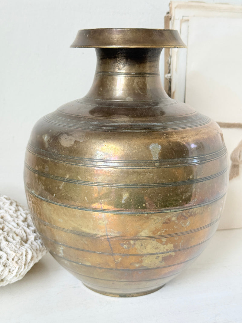 A vintage metallic bronze-colored ceramic vase with a flared rim and horizontal ridges, displayed on a white surface next to a white textured object.