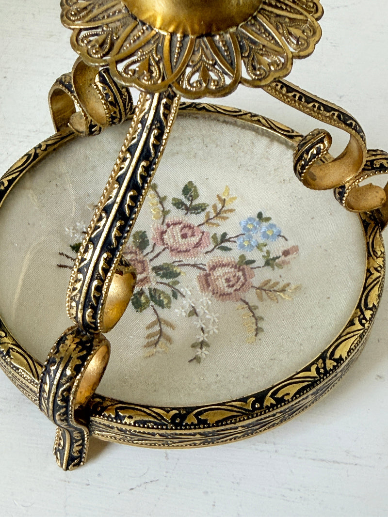 An ornate vintage-style gold and black metal wall hook with a circular glass insert featuring a delicate pink and blue floral painting.