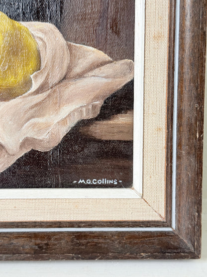 Close-up of a framed still life painting showing part of a yellow fruit, draped cream fabric, and a dark brown background, with the signature &