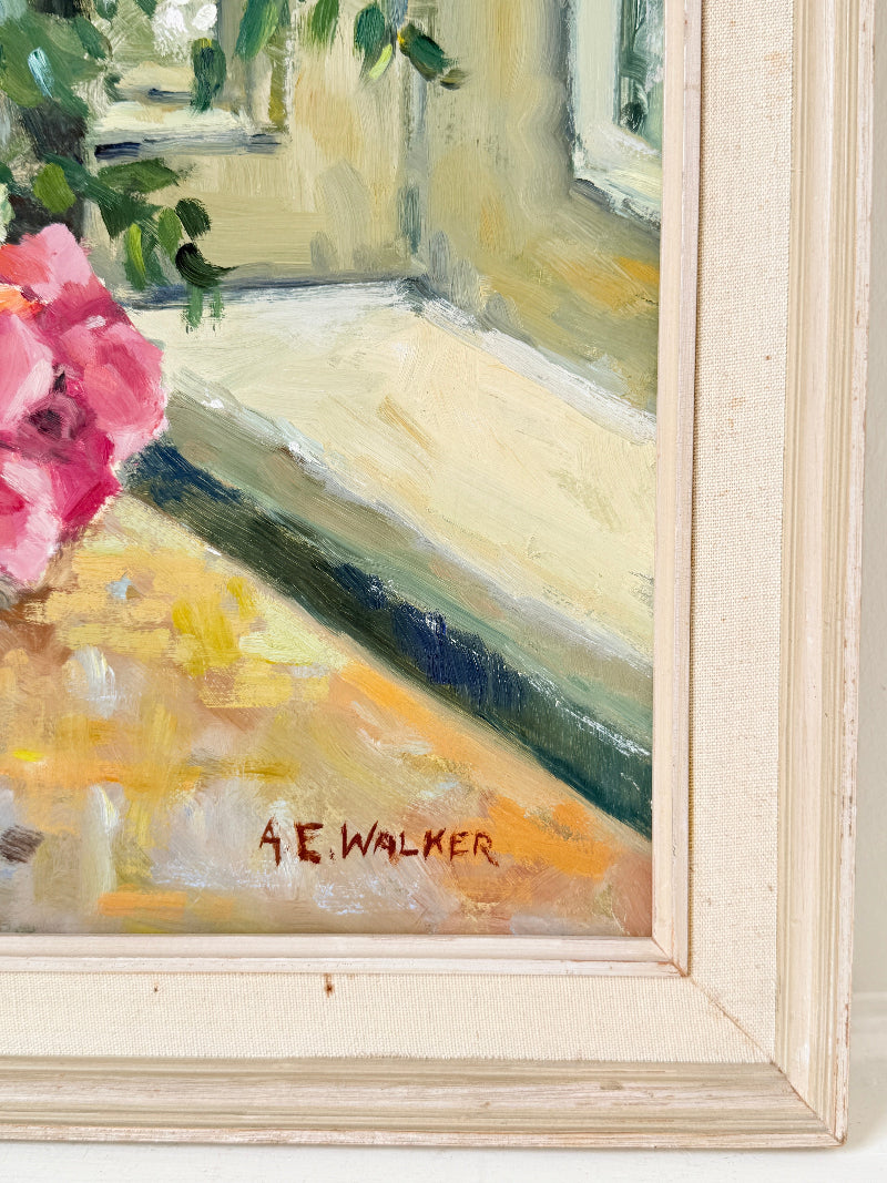 Close-up of a framed painting showing a sunlit interior corner with pink flowers at the left edge, painted in warm cream, yellow, green, and blue tones. The artist signature &