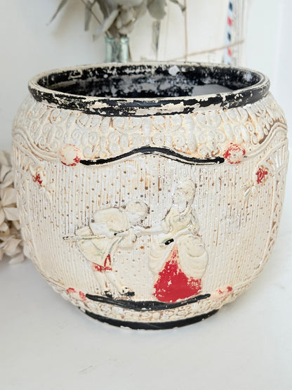 A vintage-style ceramic planter pot with a distressed off-white glaze, featuring hand-painted figures in red and black, set against a softly blurred neutral background.