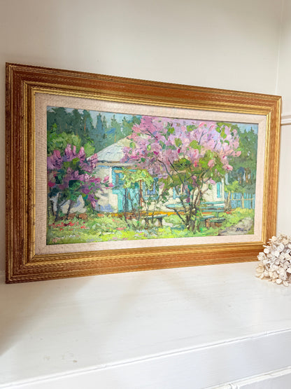 A framed landscape oil painting of a cottage garden with pink flowering trees, displayed on a white mantel. The painting features a white cottage with blue shutters, surrounded by green foliage, purple flowers, and a prominent gold frame.
