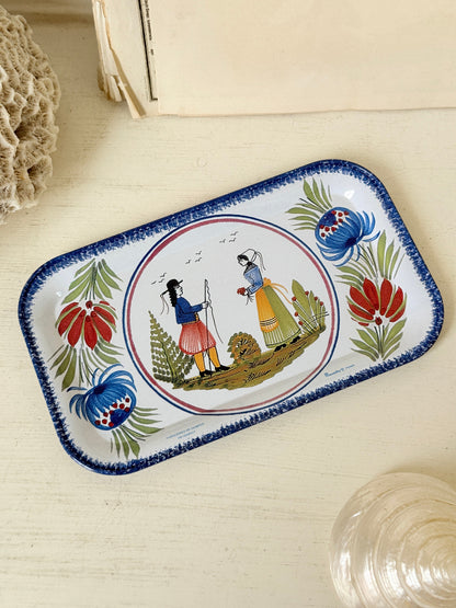 A rectangular white enamel tray with a blue hand-painted border and a central illustration of two figures in traditional clothing, surrounded by floral motifs, displayed on a light wooden surface.