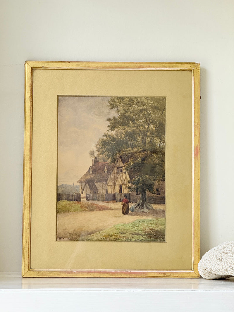 Antique framed watercolour of English village street with thatched cottages and figures, in gilt frame with wide mount, on white surface.