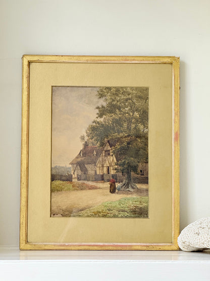 Antique framed watercolour of English village street with thatched cottages and figures, in gilt frame with wide mount, on white surface.