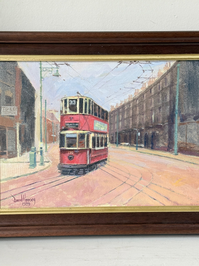 A framed painting depicting a red double-decker street tram traveling down a city street, housed in a dark brown wooden frame with a gold inner border.