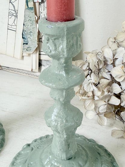A close-up of a distressed sage green ceramic candlestick holder with a red pillar candle inserted, set against a styled background with dried flowers and vintage papers.
