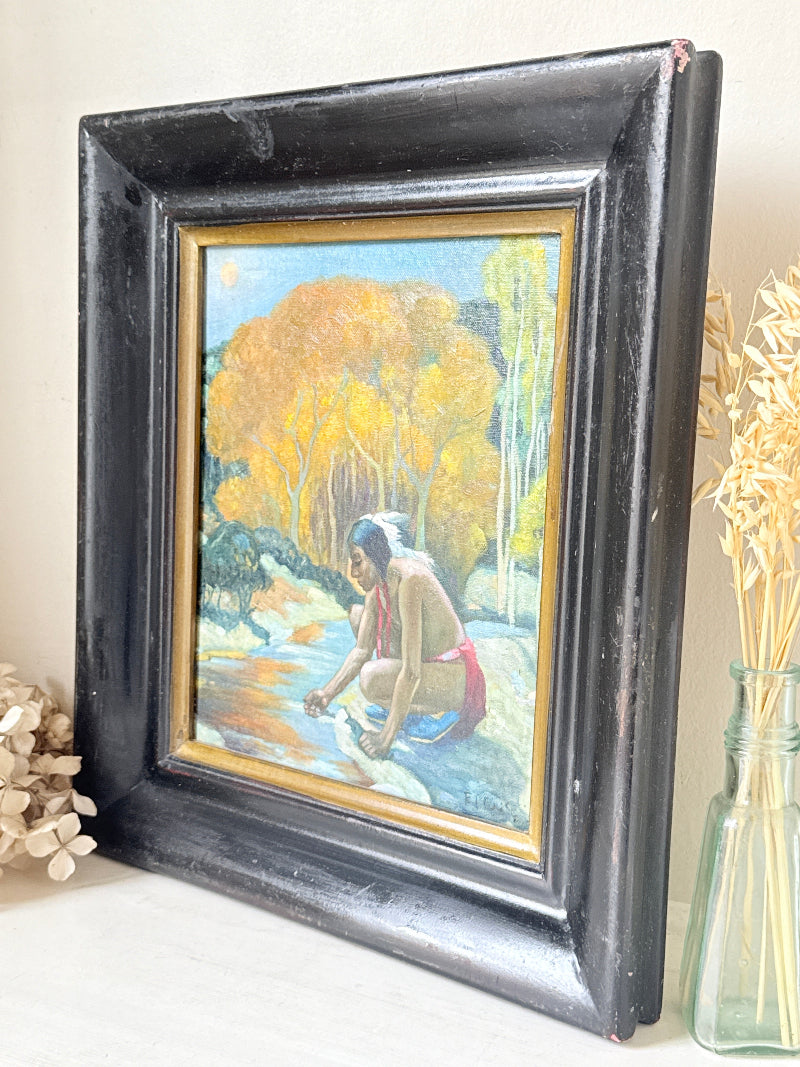 A framed vintage painting depicting a Native American woman by a river, set in a distressed black frame with a gold inner border. The painting features yellow autumn trees and a blue sky, displayed on a shelf next to dried flowers in a glass vase.