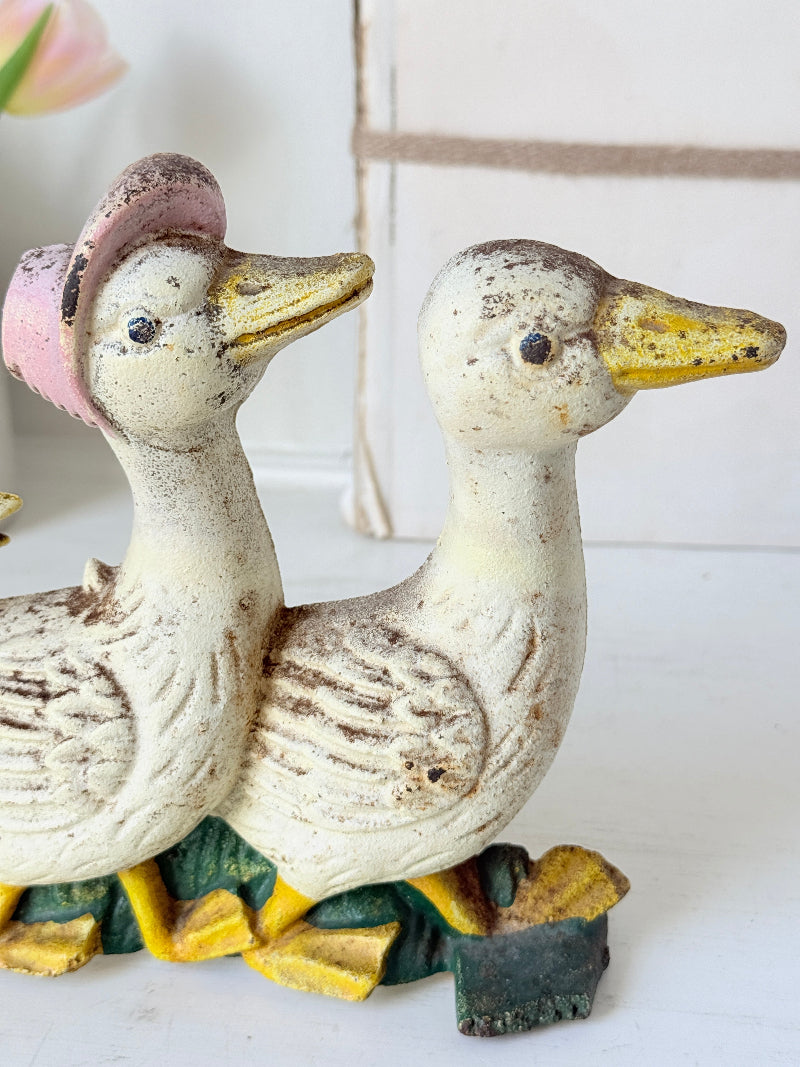 A pair of vintage ceramic duck figurines with distressed cream bodies, yellow beaks and feet, one wearing a pink hat, set against a softly blurred white background.
