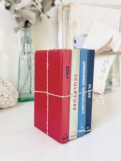 Four small hardcover books tied together with twine, standing upright on a white surface. The spines are red, cream, light blue, and dark blue, with white text reading &