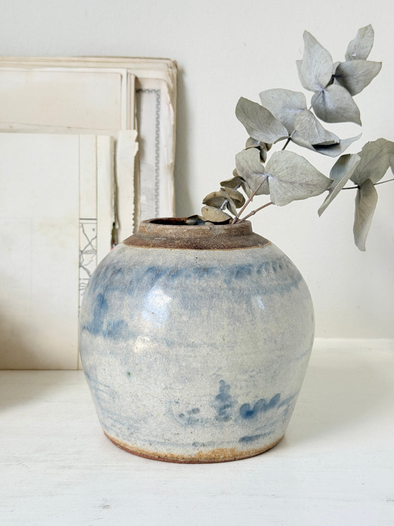 A handmade ceramic vase with a blue and white marbled glaze and a rustic brown rim, holding a dried eucalyptus branch, set against a neutral background with a vintage picture frame.