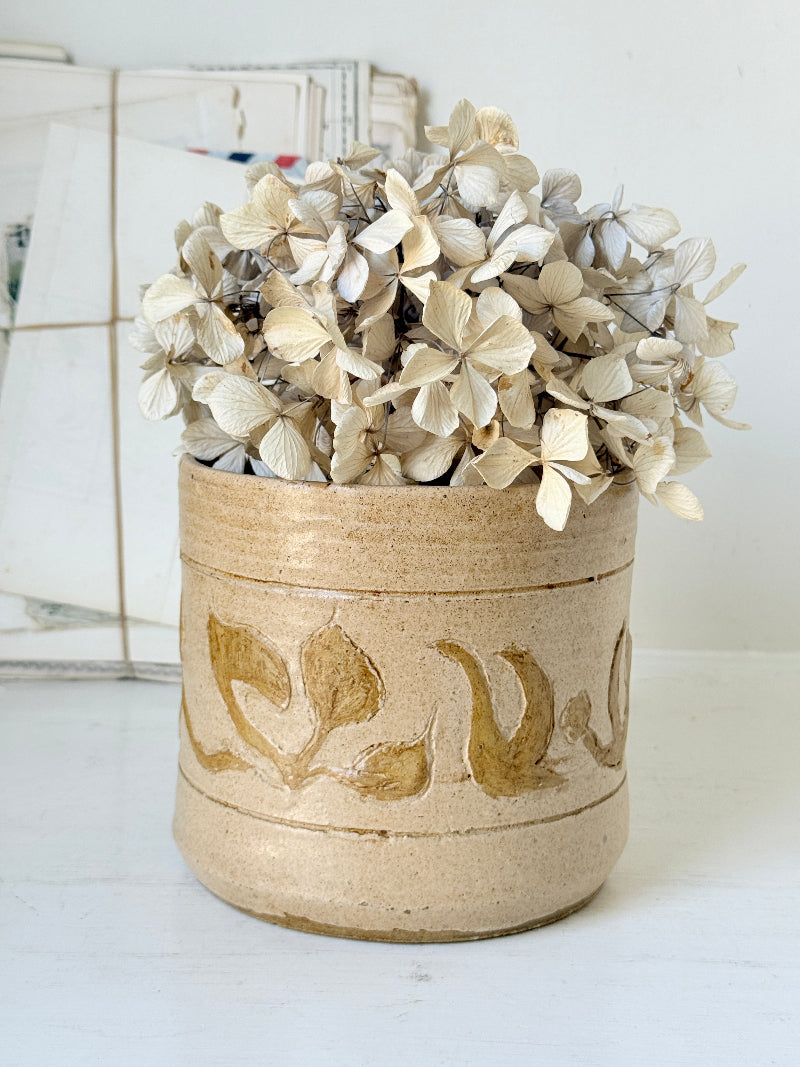 A handmade ceramic pot with a textured beige glaze and gold leaf leaf patterns, filled with dried white hydrangeas, set against a soft, light background.