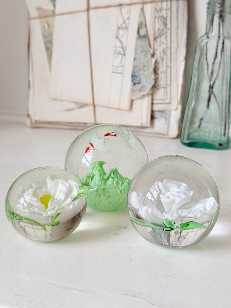 Trio of white flower glass paperweights in graduated sizes with green leaves, photographed on a white surface