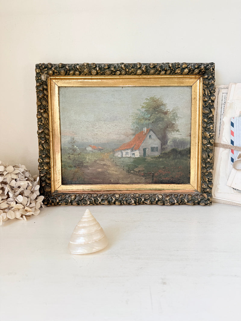 A framed vintage landscape painting of a small white house with a red roof, displayed on a white surface with decorative props.